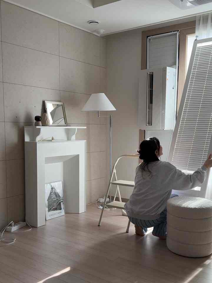 ナチュラルな色調のリビング、白い壁暖炉コンソールとフロアランプ、窓用エアコンとブラインドが配置された雰囲気のあるインテリア