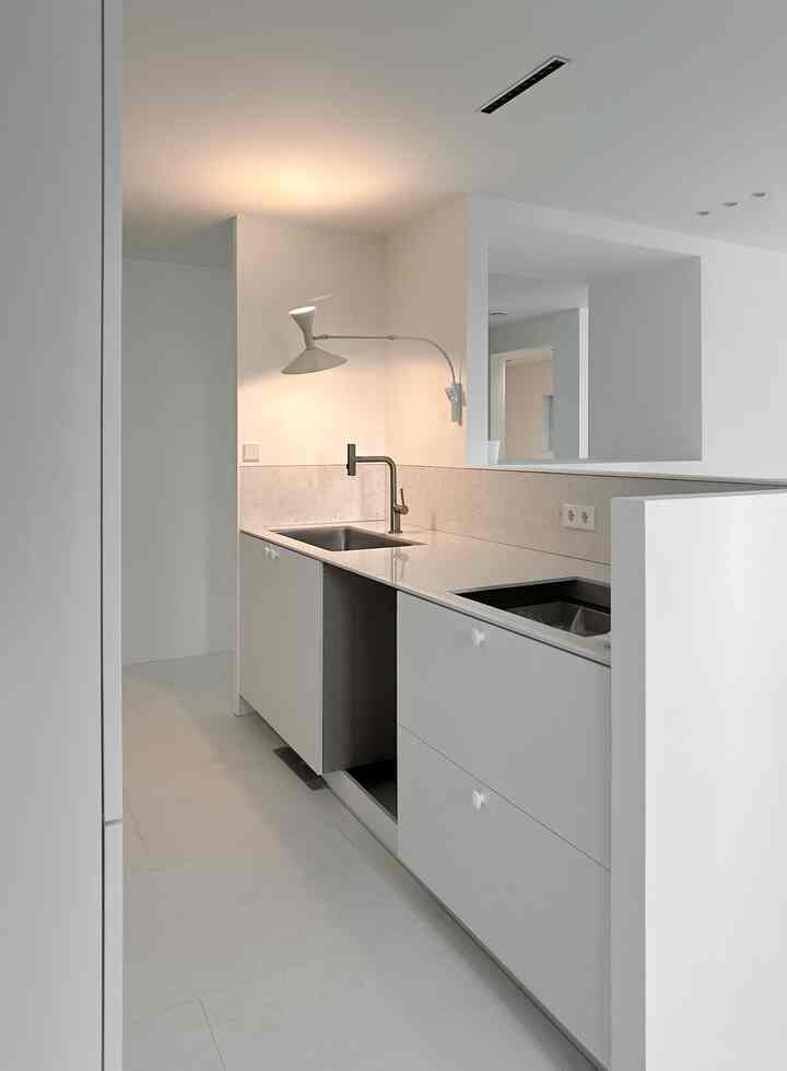 清潔感のあるミニマルな白いキッチンスペース、壁付けライトとエンジニアードストーンのカウンターが配置されたインテリア