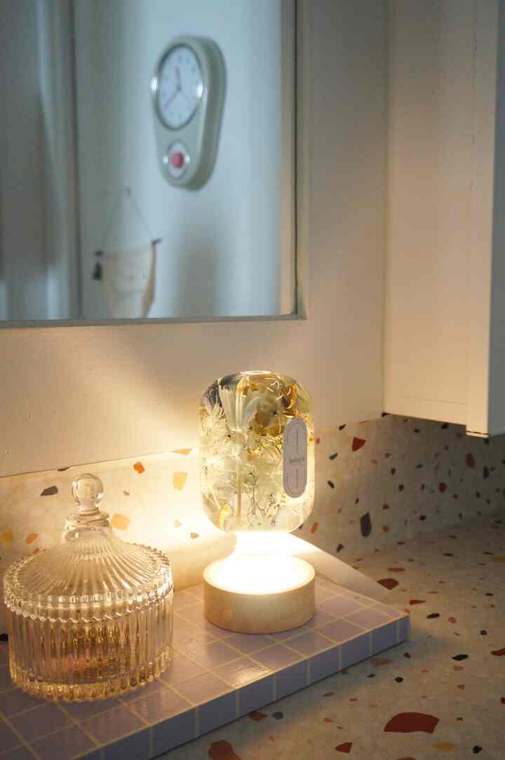 白い壁のキッチンコーナー、ベージュのタイルの上に花柄のテーブルランプとガラス容器が置かれたナチュラルな空間