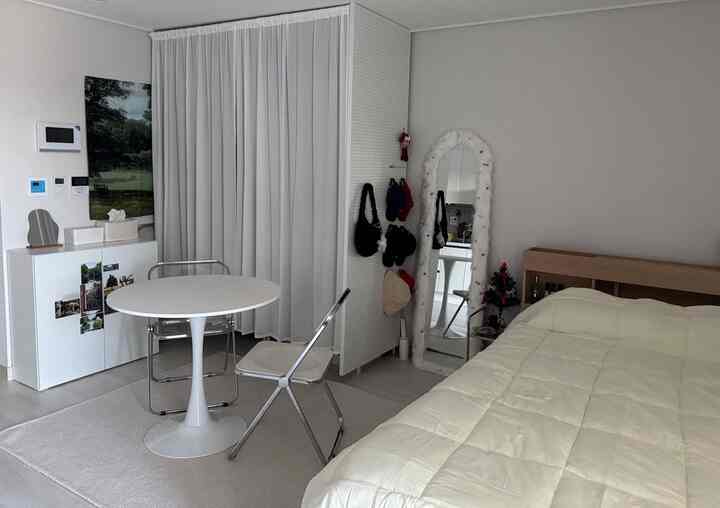 明るい白を基調としたワンルーム寝室、シンプルな丸テーブルと折りたたみ金属椅子、クローゼット仕切り壁と全身鏡が調和した印象的な空間