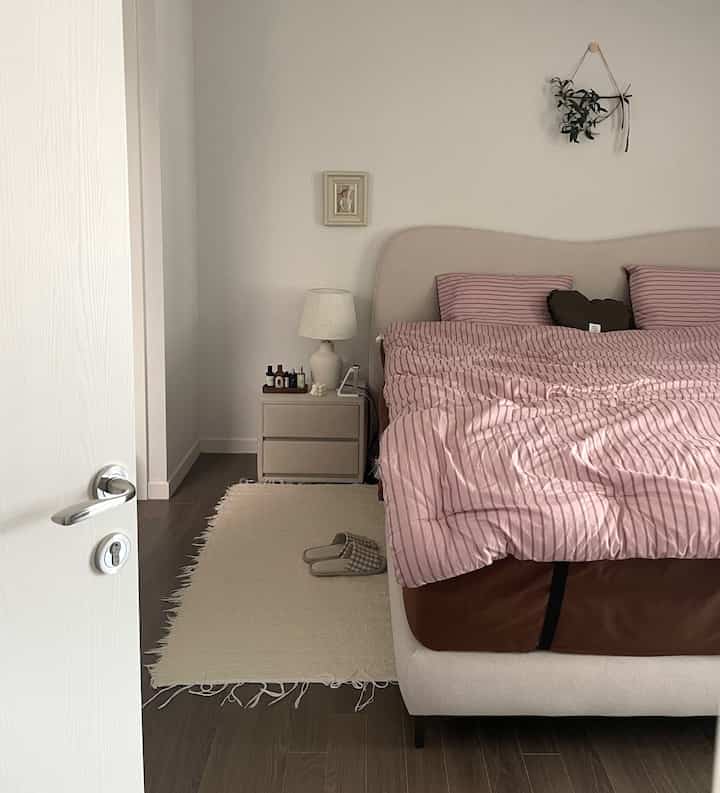 白とベージュの色調の寝室、ピンクのストライプ寝具とナイトテーブルの装飾が落ち着いた雰囲気のインテリア