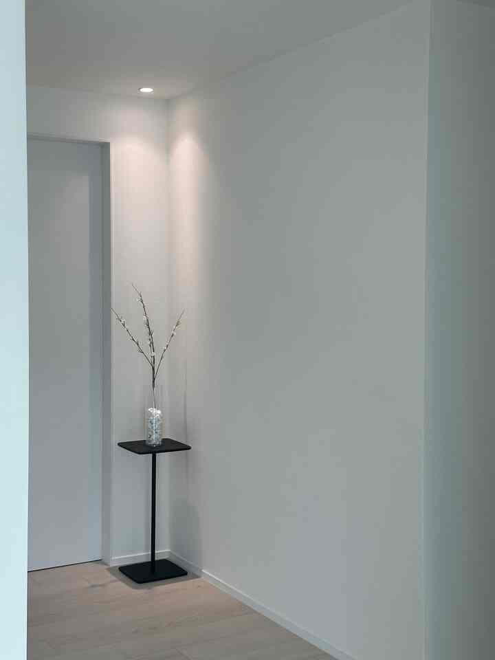 白と木目調の床が調和した玄関、白い壁と黒い小さなスタンドに花瓶が配置されたミニマルなインテリア