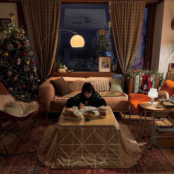 暖かいベージュの色調のリビングルーム、中央に折りたたみこたつテーブルがあり、毛布とオレンジの椅子が落ち着いた雰囲気で配置されている