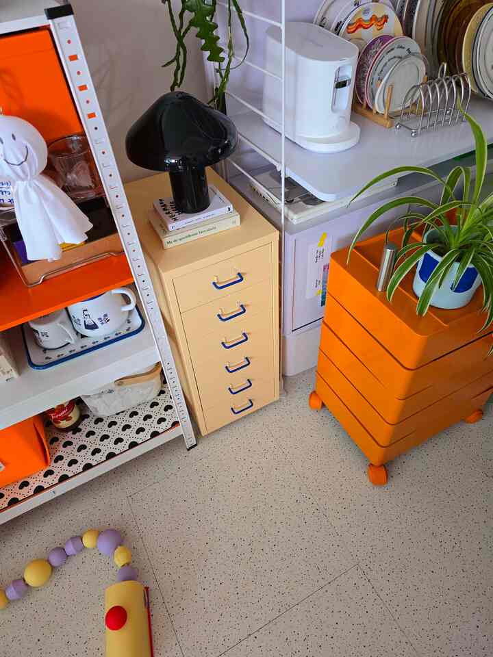 ベージュとオレンジの収納が配置された小さなキッチン兼多目的スペース、植物とテーブルランプがアクセントの温かみのある空間