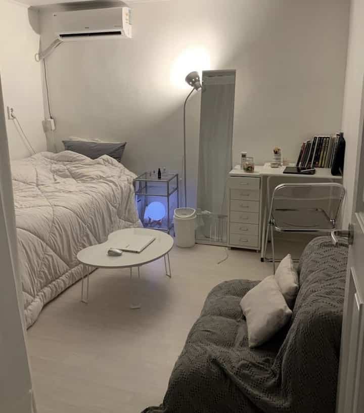 White-toned studio bedroom featuring a lift-up bed, sofa, and full-length mirror in a simple, cozy setup
