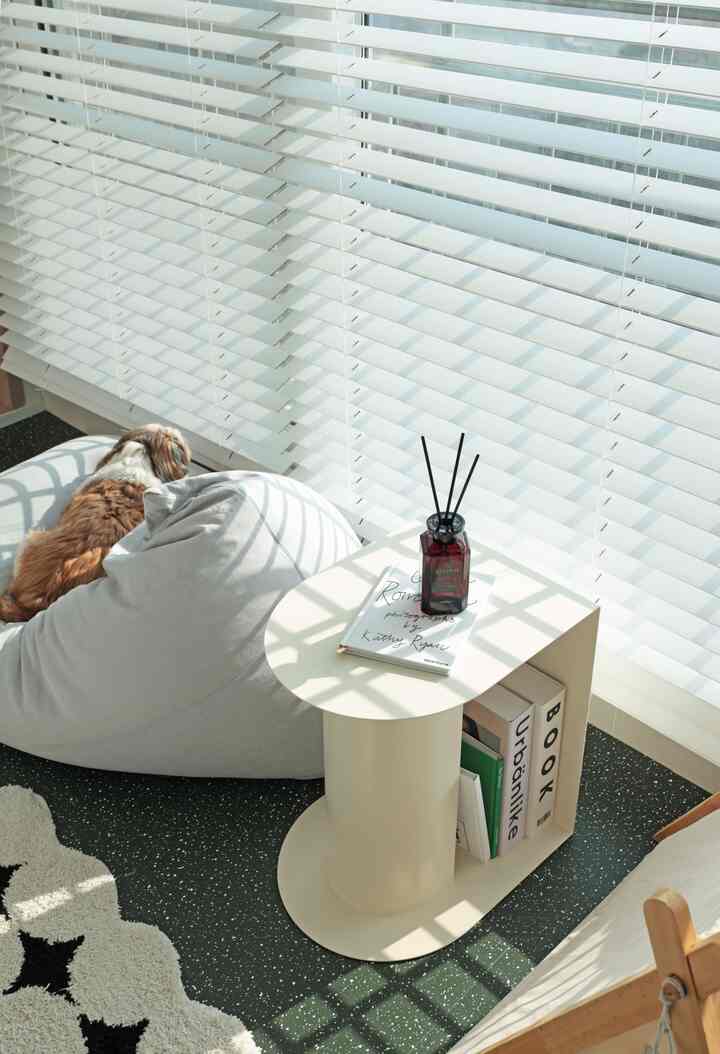 明るいベージュの半円形サイドテーブルとグレーのビーズクッション、窓際に白いブラインドがある雰囲気のあるベランダ