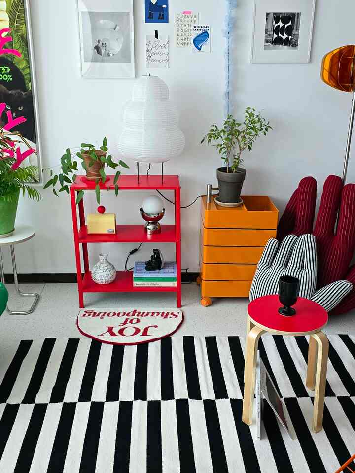 白い壁と床を背景に、赤いキャスター付きシェルフとストライプのラグ、さまざまな照明と植物が配置されたミッドセンチュリーモダンのリビング空间