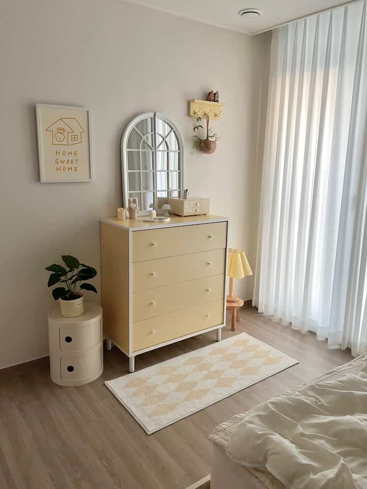 やわらかな黄色とアイボリーの色調でまとめられた寝室の一角、バターイエローのチェストと棚、温かみのある照明が配置された印象的なインテリア