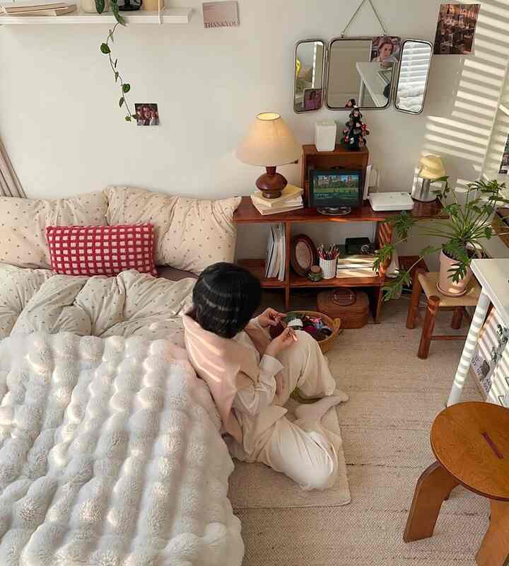 白い壁とベージュのナチュラルな色調のワンルーム寝室、低めのベッドと木目調の棚、暖かい照明と植物が配置された居心地の良いインテリア