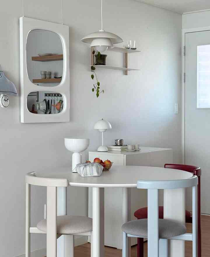 明るいホワイトトーンのダイニング、丸テーブルと異なる3色の椅子、壁に鏡と棚が配置された印象的な空間