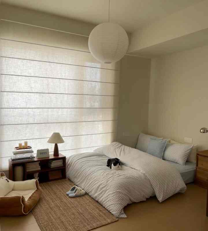 柔らかいベージュと木目調のトーンが調和する寝室、ストライプの寝具と木製ナイトテーブル、低いマットレスの上に黒白の猫が横たわる暖かみのある空間