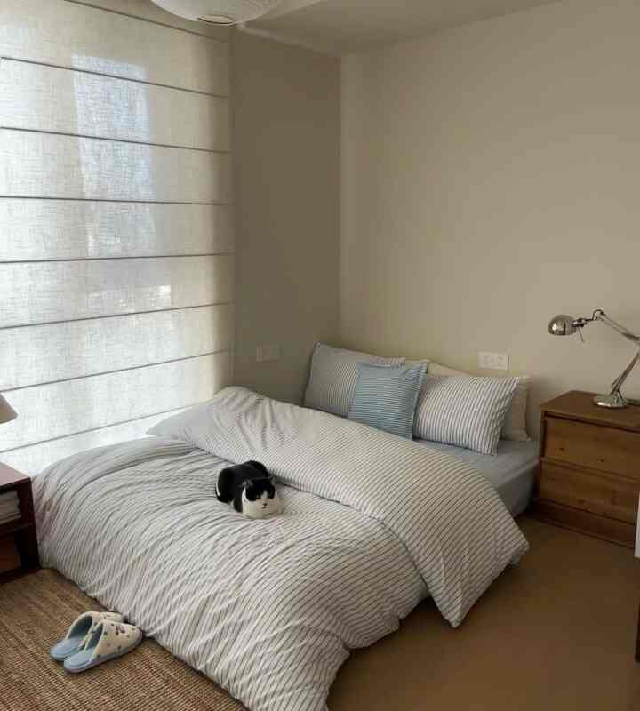 ナチュラルな色調の寝室、ストライプ柄の掛け布団と木製ナイトテーブル、ベッドの上に猫が座っている印象的な空間