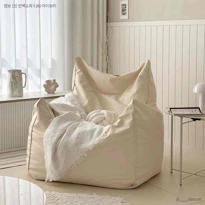 ナチュラルなベージュ色調の小さな寝室、中央にベージュのビーズクッションソファと白いブランケットが配置された明るく居心地の良い雰囲気のインテリア