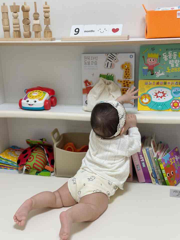 ホワイトとナチュラルトーンの子供部屋、赤ちゃんがおもちゃ棚に手を伸ばす印象的で温かみのある空間