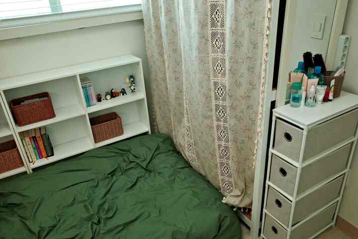 白を基調とした内装のナチュラルな小さな寝室、緑のベッドカバーと花柄のカーテンが配置された清潔感のあるインテリア