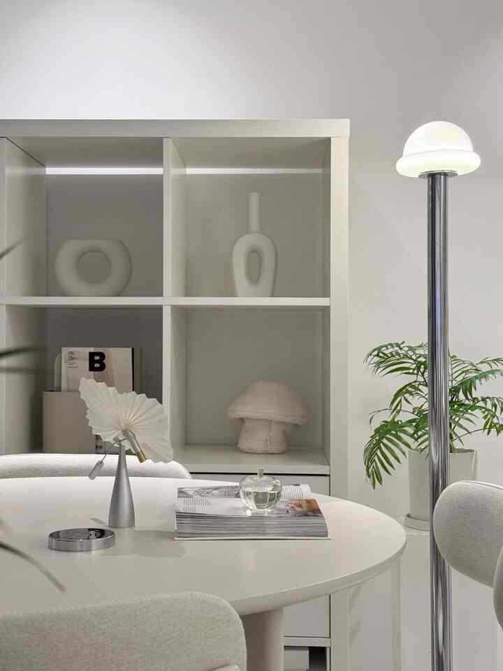 Bright white-toned living room featuring a round dining table with chairs, floor lamp, bookshelf, and plant in a simple setting