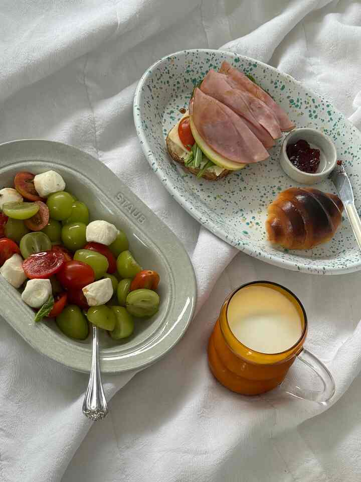 白色調の寝室に楕円形の皿とフルーツ皿、ブラウンの耐熱グラスが置かれた居心地の良いベッドでの食事シーン