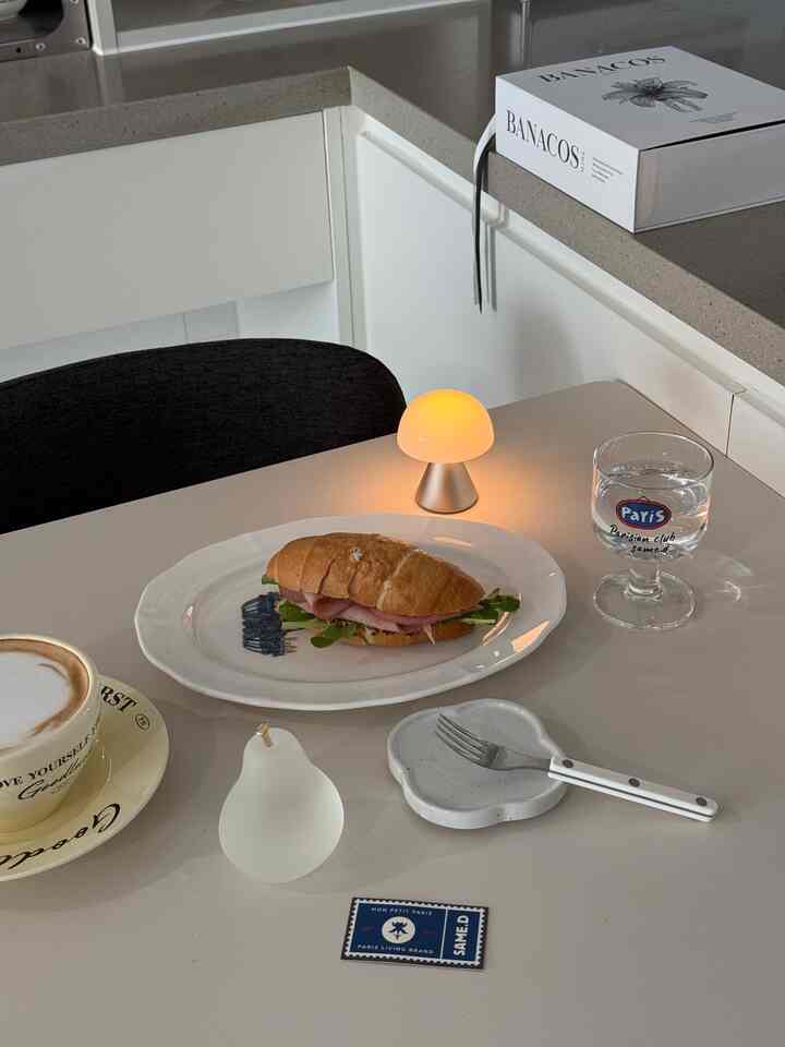 White and gray toned kitchen dining area featuring a salt bread sandwich, coffee cup, water glass, and mushroom mood lamp, creating a cozy home cafe vibe