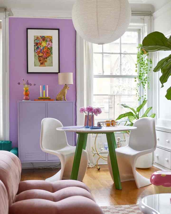 A lavender-accented living room featuring white chairs around a round table, plants near the window, and a hanging pendant light creating a cozy bright interior