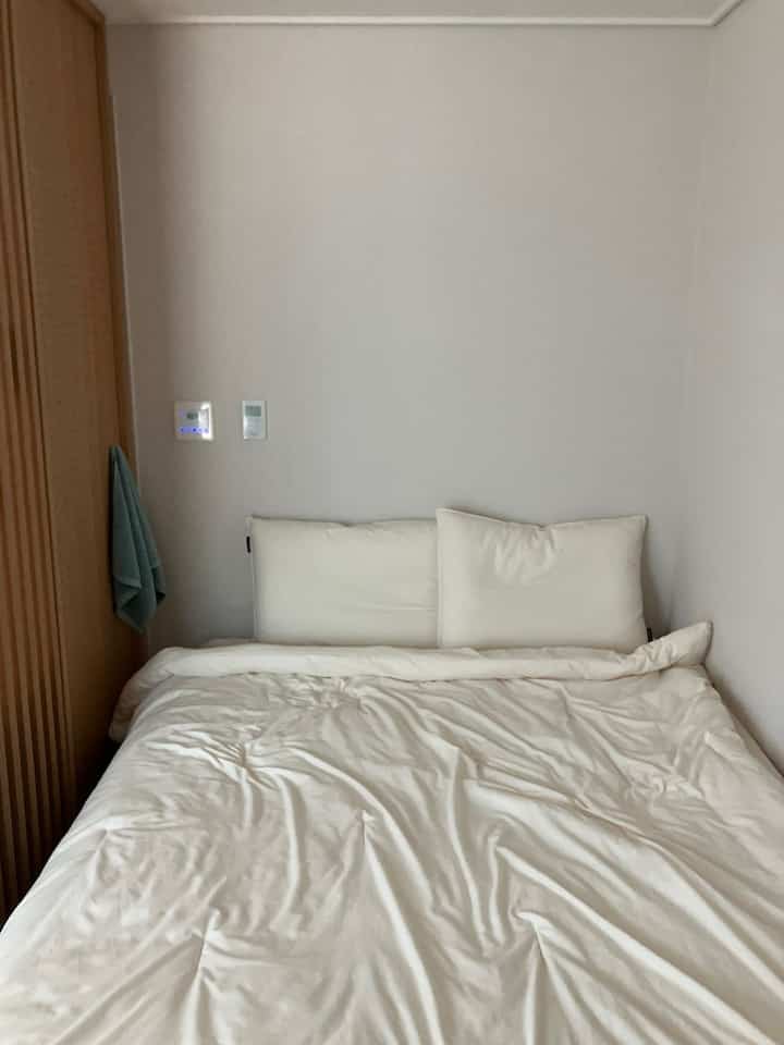 白を基調とした寝室、クイーンサイズのベッドと間仕切りの隣に掛けられたタオルが配置されたシンプルで清潔感のあるインテリア