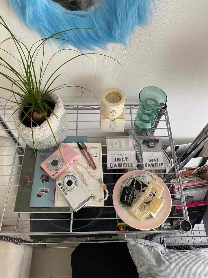 青いふわふわの鏡が壁に掛かり、金属製の収納ラックに植物や小物が整然と置かれた、小さな一人暮らし用6畳部屋のインテリア