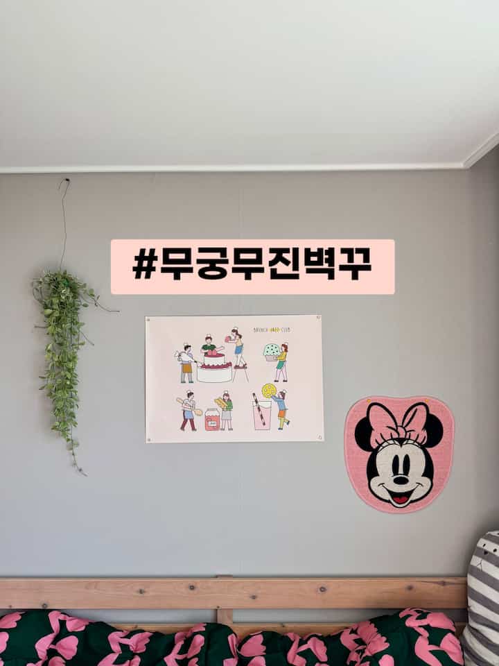 グレーの壁と木目調のベッドフレームがある子供部屋、ピンクの寝具と壁飾り、壁に掛かったグリーンの植物が調和した温かみのある空間