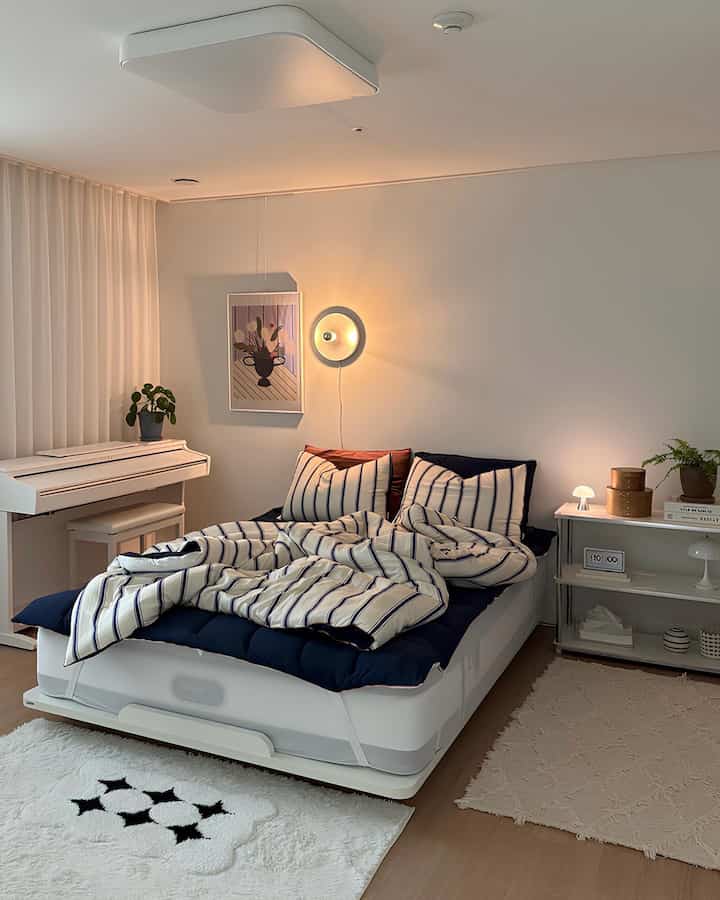 A white and navy toned bedroom featuring a central bed with striped bedding, digital piano, lighting fixtures, and shelving creating a cozy atmosphere
