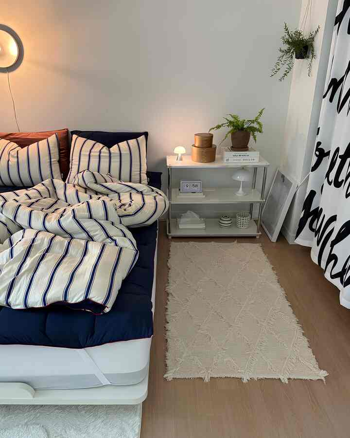 ネイビーとホワイトの色調の寝室、ストライプの寝具と木目調の床、ナイトテーブルに置かれた照明と植物が配置されたシンプルで清潔感のあるインテリア