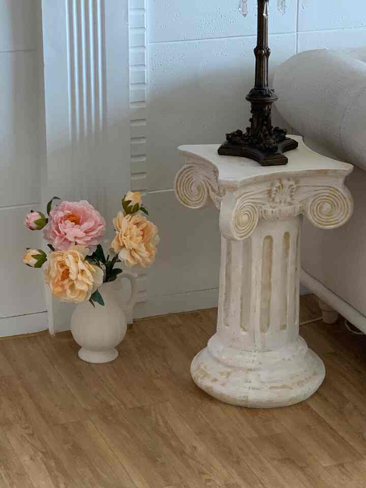 白い壁と木目調の床、アンティーク調の柱台、造花の飾られたリビングコーナーのインテリア