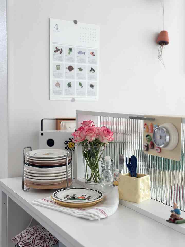 明るいホワイトトーンのキッチン棚にイケアの皿が整然と積まれ、中央にはピンクのバラが活けられた花瓶が置かれた、清潔感のあるナチュラルなインテリア