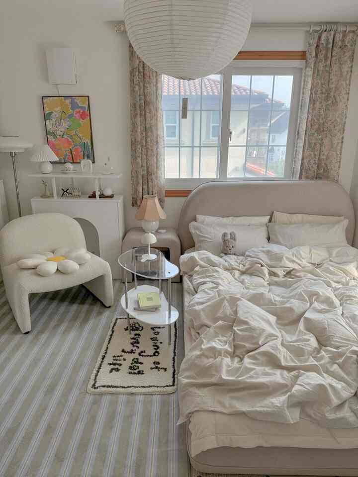 ベージュと白のトーンの寝室、柔らかいベッドとフロアアームチェア、花柄カーテンが配置された雰囲気のあるインテリア