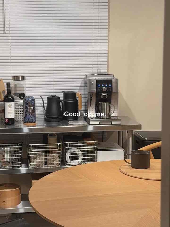 白いブラインドと木目調の丸テーブルがあるキッチン空間、ステンレス作業台にコーヒーマシンと道具が配置された雰囲気のあるインテリア