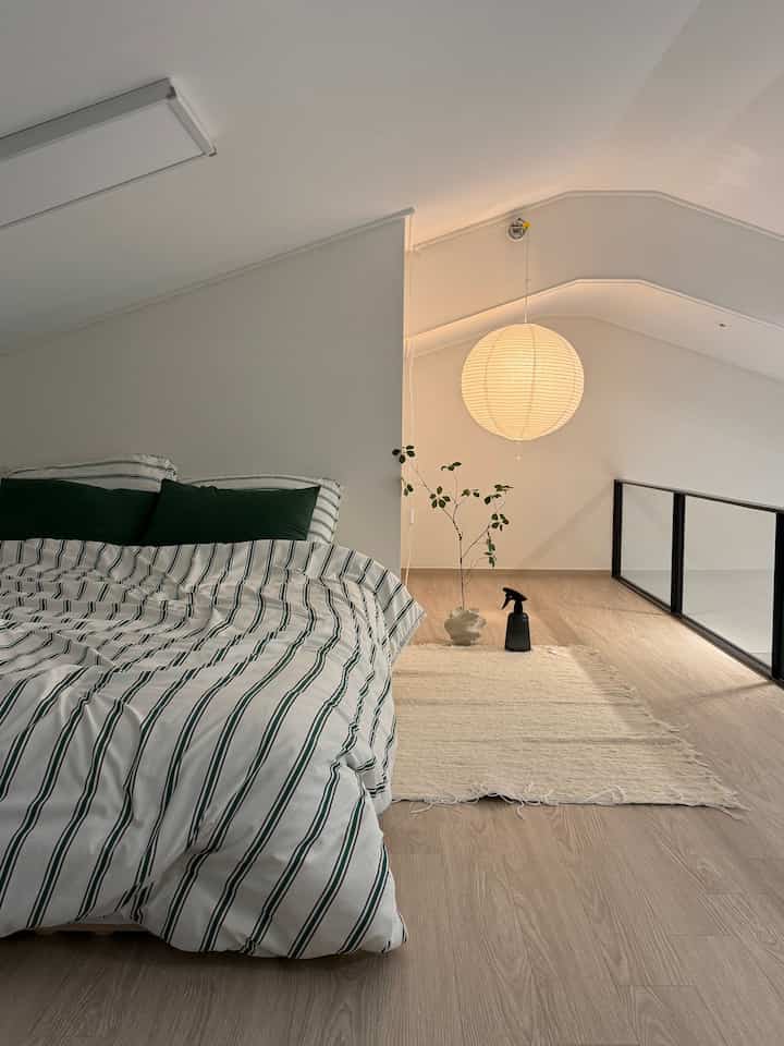 ナチュラルカラーとホワイトトーンのロフト寝室、ストライプの寝具とペンダントライト、植物が配置された雰囲気のある空間