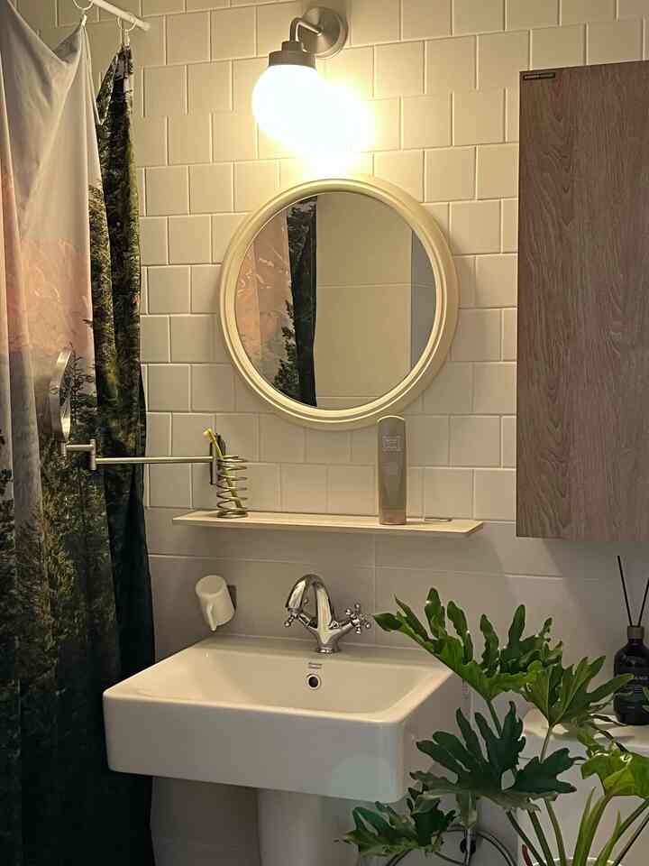 白いタイルの壁と木目調のキャビネットが調和したバスルーム、丸い鏡と照明、植物が配置された雰囲気のある空間