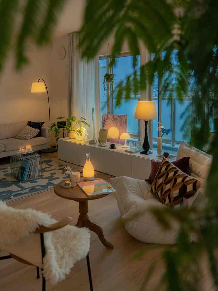 Bright and warm living room with white walls and curtains, soft wood tone flooring, cozy sofas, and ambient lamps creating a relaxed atmosphere