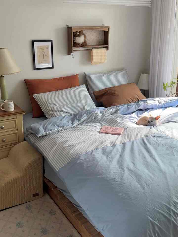 柔らかなブルーとブラウンのトーンの寝室、低い木製ベッドと多彩な枕が置かれた温かみのある居心地の良い空間、小さな犬がベッドの上でくつろいでいる