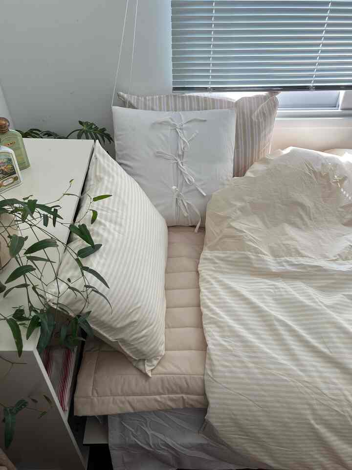 明るいホワイトとベージュの寝具と枕、植物が置かれた居心地の良い寝室、窓にはシルバーのアルミブラインドがかかる空間