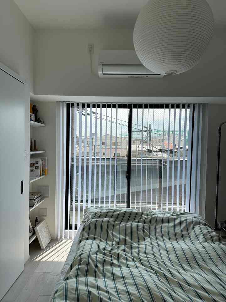 ライトグレーと白の寝室、ベランダとバーチカルブラインドが配置されたすっきりとした空間