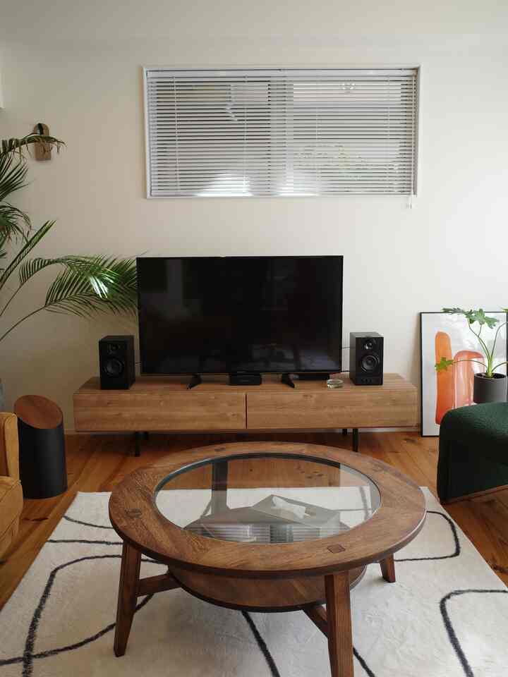 木目調と白を基調とした1LDKのリビング、丸い木製のコーヒーテーブルとテレビ台、植物とポスターが配置されたシンプルなインテリア