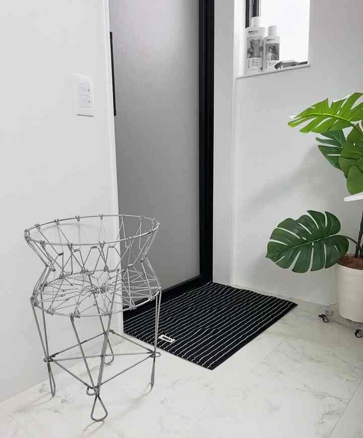 白と黒のトーンのバスルーム、金属製ランドリーバスケットとストライプのバスマット、観葉植物が配置された印象的な空間