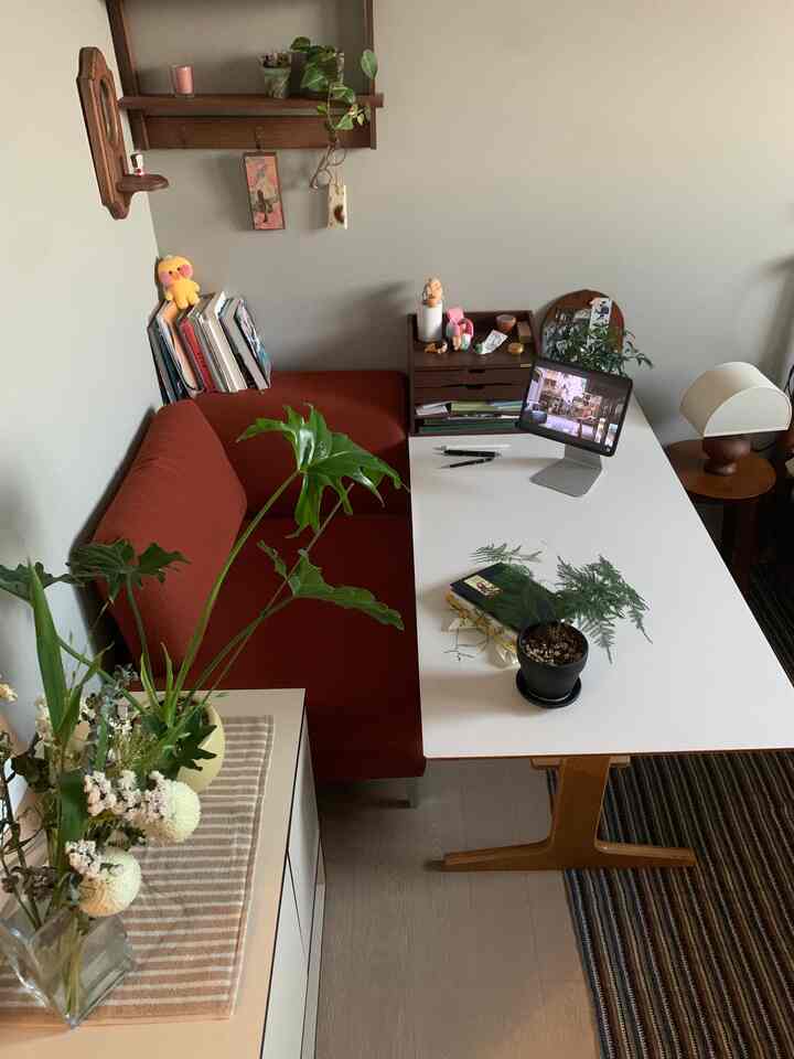ナチュラルな色調のリビング、赤みのあるソファと白いデスクが配置された植物豊かな落ち着いた空間