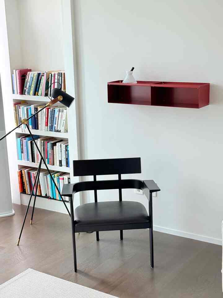 Modern living room with natural tone flooring and white walls, featuring a black armchair, black floor lamp, and red wall-mounted shelf