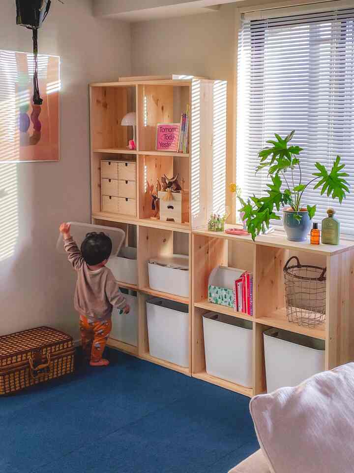 ナチュラルな色調の子供部屋、木製棚が配置され、青いカーペットで遊ぶ子供がいる温かみのある空間