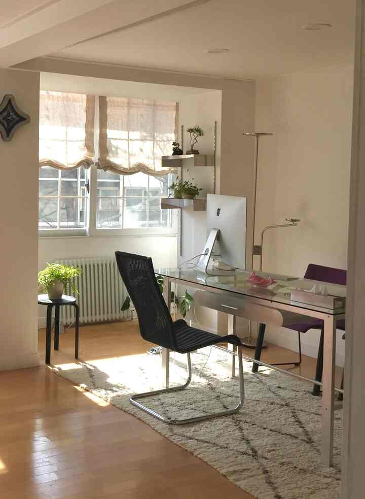 明るいベージュと白を基調としたワンルームのホームオフィス、ガラスデスクとオフィスチェア、植物が配置されたミニマルなインテリア