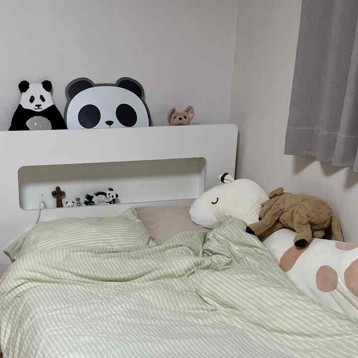 白い壁とベージュのカーテンがある寝室、ストライプの掛け布団カバーと複数の動物ぬいぐるみが配置された雰囲気のある空間