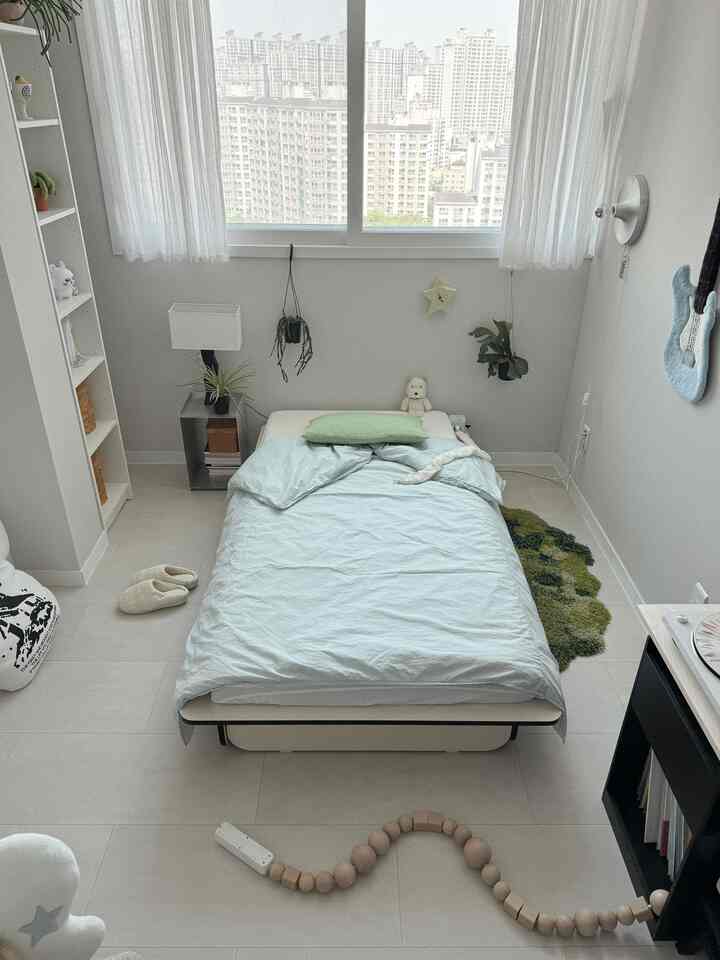 ホワイトとグリーンのトーンの寝室、中央にシングルベッドと緑の枕、植物とホワイトカーテンが調和したシンプルでナチュラルなインテリア