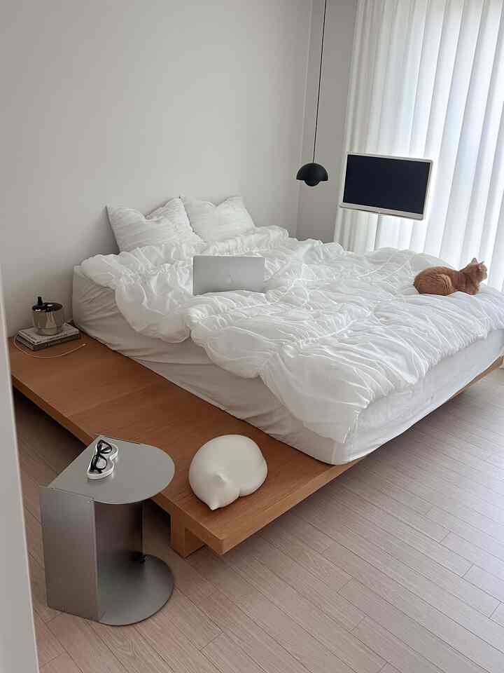 A modern, simple bedroom featuring white and wood tones, with a wooden platform bed, side table, and a cat resting on the bed.