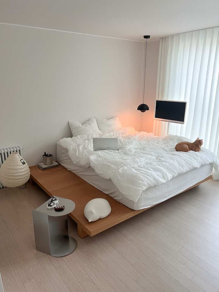 Simple bedroom in white and natural tones featuring a wooden bed with plush bedding, a cat on the right side, and pendant lighting