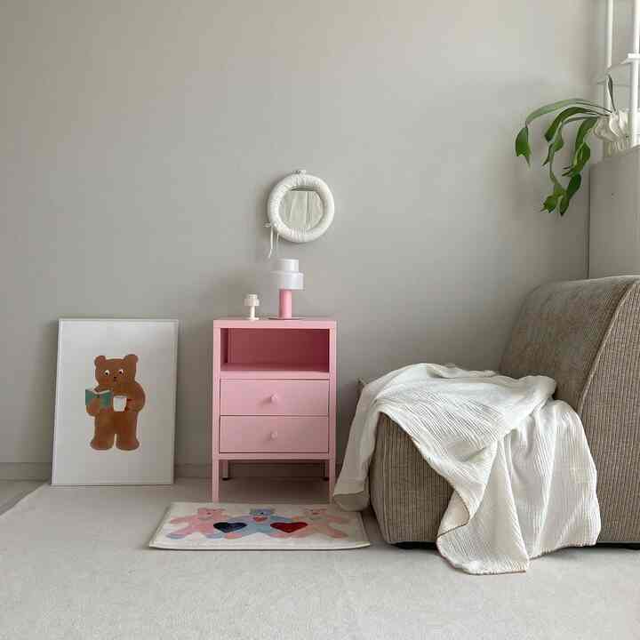 白とピンクのトーンのリビング、ピンクの小さなチェストとベージュのアームチェア、ブランケットで温かみのあるミニマルな印象のインテリア