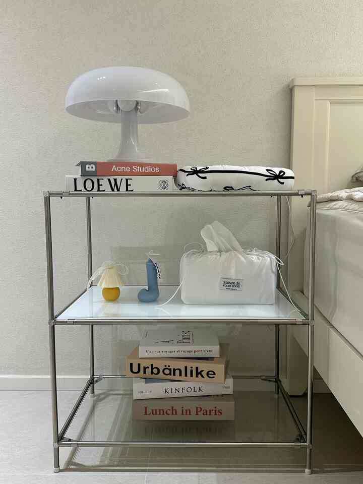 白を基調としたモダンな寝室、金属フレームの3段棚ナイトテーブルにランプや本、ティッシュケースが配置された雰囲気のあるインテリア